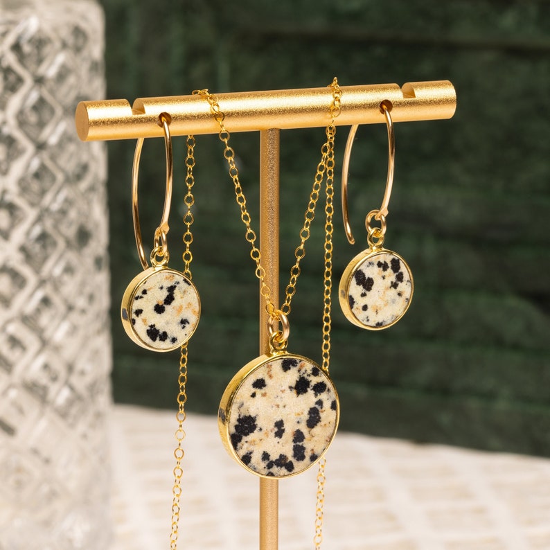May include: A gold necklace and earrings set with round dalmatian jasper stone pendants. The stones have a white and black speckled pattern.