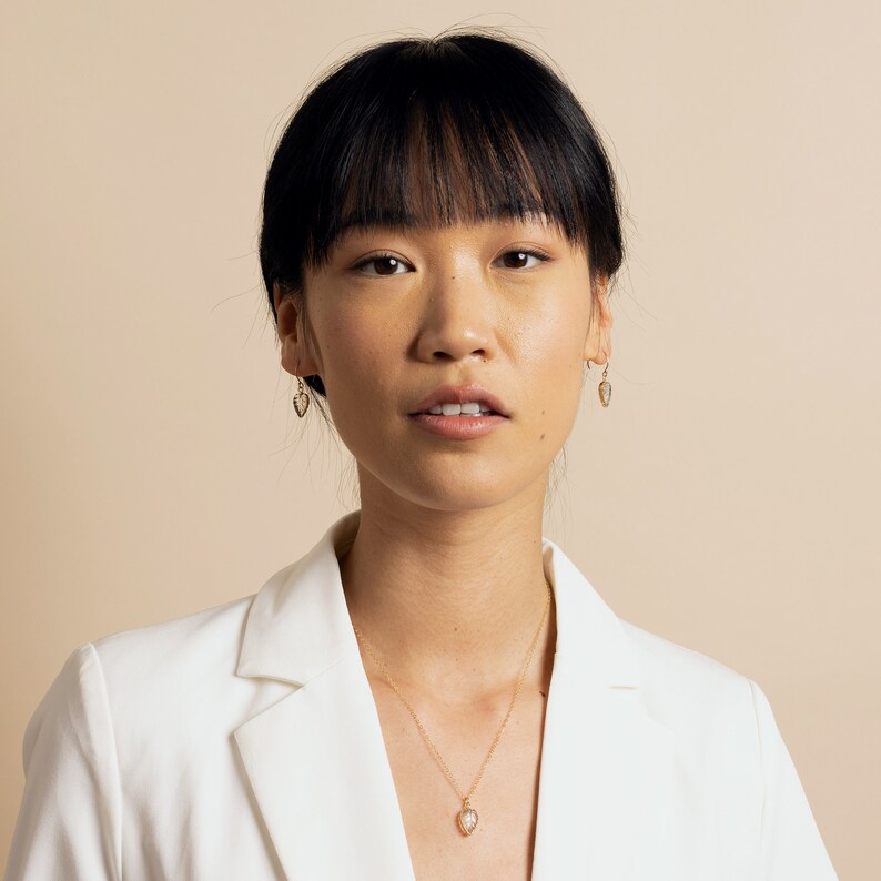 May include: A woman with dark hair wearing a white blazer and a gold necklace with a small heart pendant. She is also wearing gold earrings with small heart pendants.