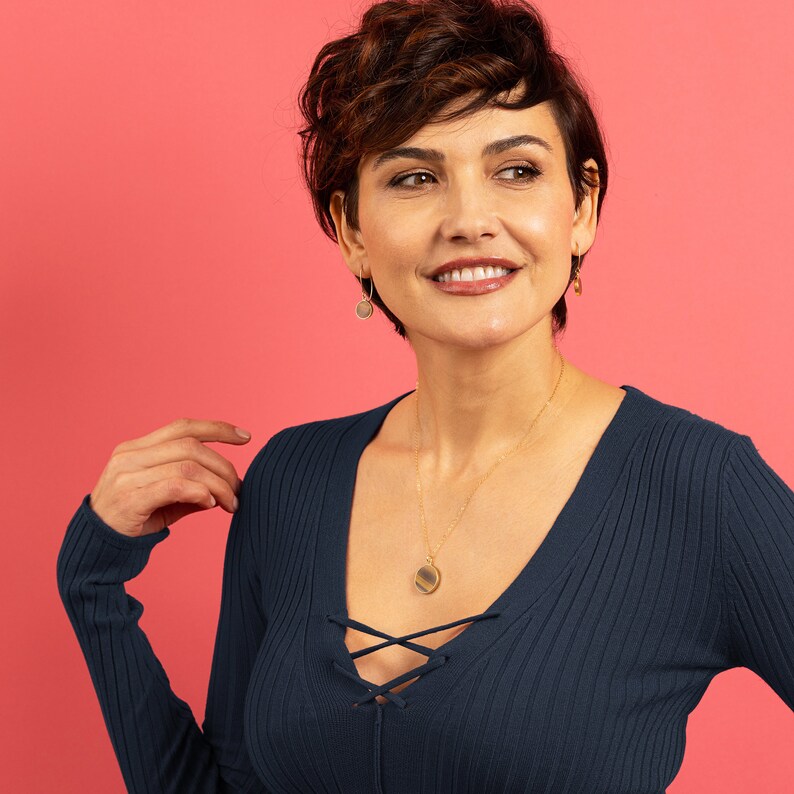 May include: A woman with short brown hair is wearing a dark blue sweater with a lace-up neckline. She is wearing a gold necklace with a round pendant and gold earrings with round pendants.