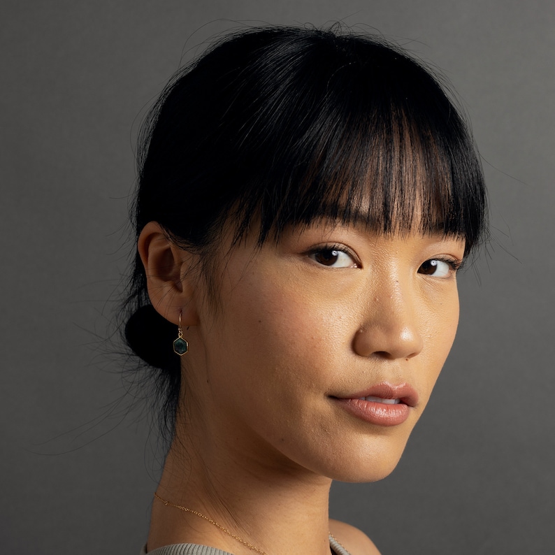 May include: A woman with dark hair and bangs wearing a gold earring with a dark green hexagonal stone.