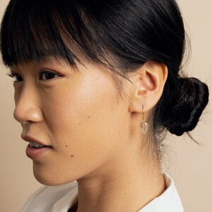 May include: A woman with dark hair is wearing a pair of gold leaf earrings. The earrings are delicate and feature a small, clear leaf charm.