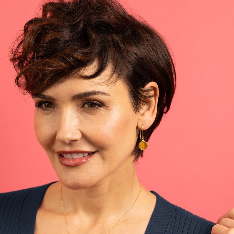 May include: A woman with short, curly brown hair is wearing gold hoop earrings with a round, textured charm. She is wearing a dark blue sweater and a gold necklace.