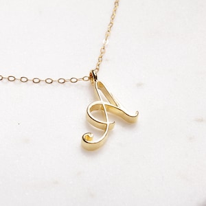 May include: A gold-colored necklace featuring a cursive letter "A" pendant. The pendant is suspended from a delicate chain. The necklace is displayed on a white marble surface, creating a simple and elegant aesthetic.