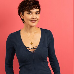 May include: A woman with short brown hair is wearing a dark blue, ribbed, long-sleeved sweater with a lace-up neckline. She is wearing a gold necklace with a round pendant and gold earrings.