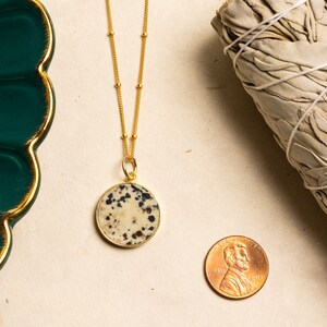 K&ouml;nnte beinhalten: Goldkette mit einem runden Dalmatiner-Jaspis-Anh&auml;nger. Der Anh&auml;nger ist hellbeige mit schwarzen Flecken. Die Kette liegt auf einer beigefarbenen Oberfl&auml;che mit einem Penny als Gr&ouml;&szlig;envergleich.