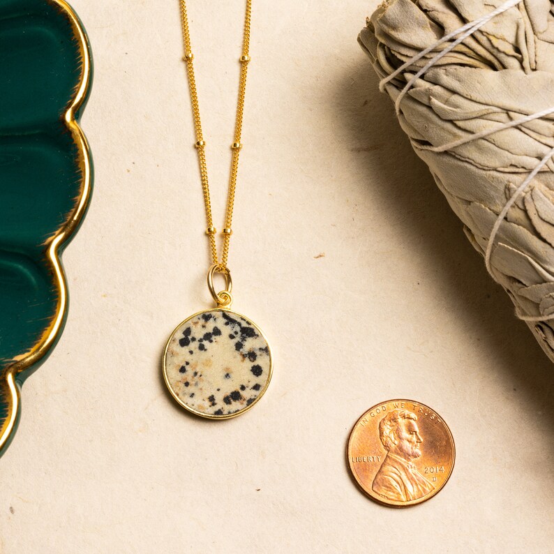 K&ouml;nnte beinhalten: Goldkette mit einem runden Dalmatiner-Jaspis-Anh&auml;nger. Der Anh&auml;nger ist hellbeige mit schwarzen Flecken. Die Kette liegt auf einer beigefarbenen Oberfl&auml;che mit einem Penny als Gr&ouml;&szlig;envergleich.