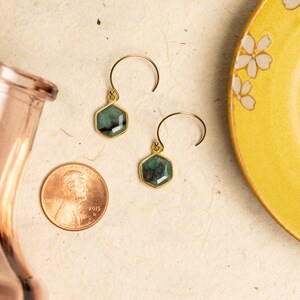 May include: A pair of gold earrings with green gemstone hexagon-shaped drops. The earrings are on a white textured surface.