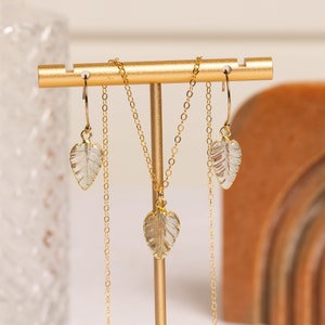 May include: A gold necklace and earrings set with clear leaf-shaped pendants. The necklace and earrings are displayed on a gold stand.