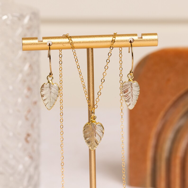 May include: A gold necklace and earrings set with clear leaf-shaped pendants. The necklace and earrings are displayed on a gold stand.