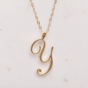 May include: A gold-colored pendant necklace featuring the cursive letter "Y". The letter is the focal point, suspended from a delicate chain. The necklace is displayed against a white background, highlighting the jewelry's design.