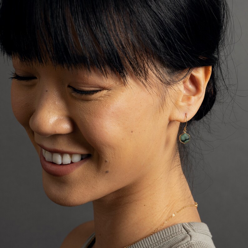May include: A woman with dark hair wearing a gold necklace and a pair of gold earrings with green gemstone accents.