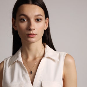 May include: A woman with long dark hair wearing a white sleeveless shirt with a collar. She is wearing a gold necklace with a black pendant.