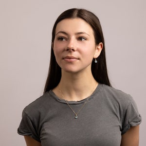 May include: A woman with long brown hair wearing a gray t-shirt and a gold necklace with a blue gemstone pendant. She is also wearing small gold earrings with blue gemstones.
