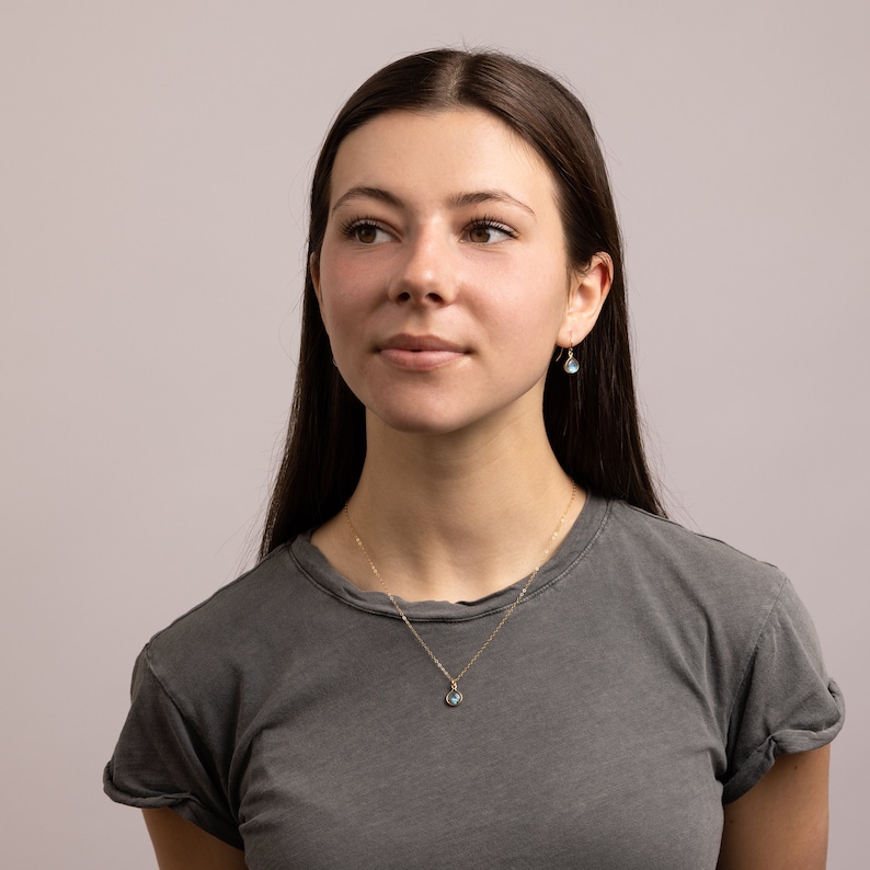 May include: A woman with long brown hair wearing a gray t-shirt and a gold necklace with a blue gemstone pendant. She is also wearing small gold earrings with blue gemstones.