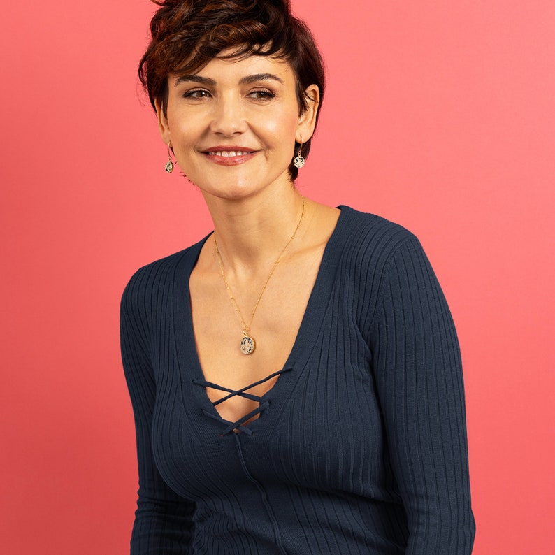 K&ouml;nnte beinhalten: Eine Frau mit kurzen braunen Haaren tr&auml;gt einen dunkelblauen, lang&auml;rmeligen V-Ausschnitt-Pullover mit einer goldenen Kettenkette und einem kleinen Anh&auml;nger. Sie tr&auml;gt auch goldene Ohrringe.