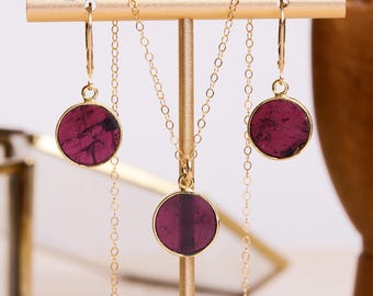 May include: A gold necklace and earrings set with round, dark purple gemstone pendants. The necklace and earrings are on a gold display stand.
