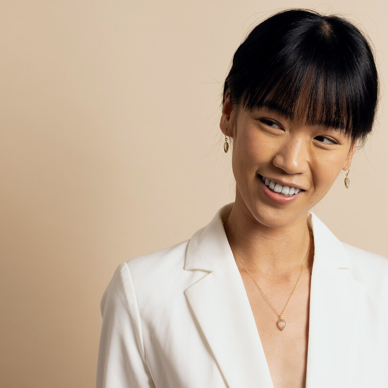 May include: A woman with dark hair and bangs is wearing a white blazer and a gold necklace with a heart pendant. She is smiling and looking to the side.