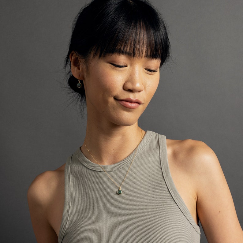 May include: A woman wearing a gray tank top with a gold chain necklace featuring a green gemstone pendant. She is also wearing small gold hoop earrings with green gemstones.