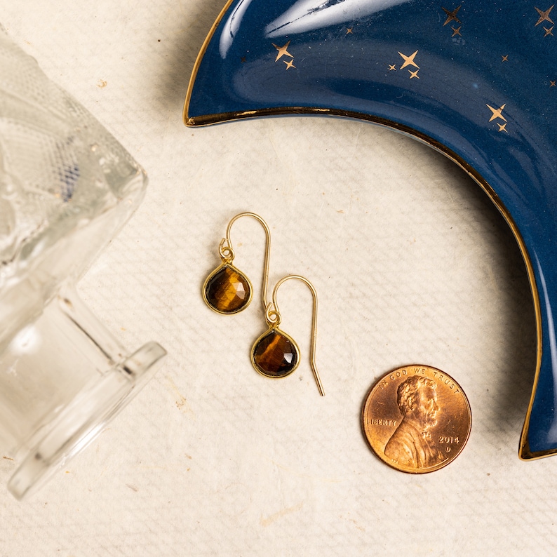 May include: A pair of gold earrings with teardrop-shaped tiger's eye stones. The earrings are shown on a white surface with a blue and gold crescent moon dish and a US penny.