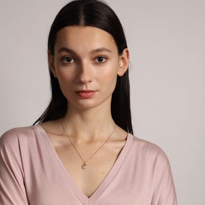 May include: A woman wearing a light pink v-neck shirt with a gold chain necklace and a small pearl pendant.