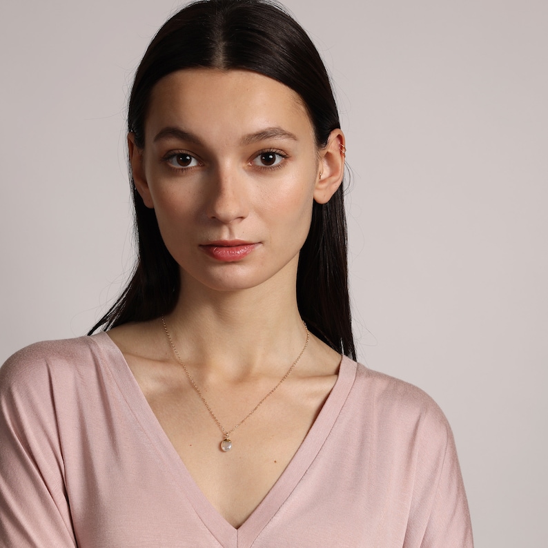 May include: A woman wearing a light pink v-neck shirt with a gold chain necklace and a small pearl pendant.