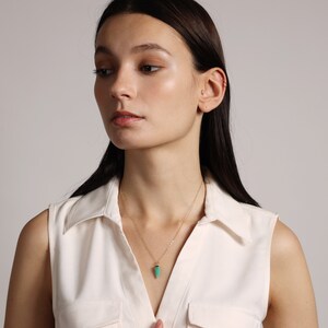 May include: A woman wearing a white sleeveless shirt with a gold chain necklace and a small, teardrop-shaped green pendant.