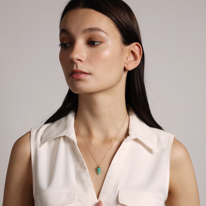 May include: A woman wearing a white sleeveless shirt with a gold chain necklace and a small, teardrop-shaped green pendant.
