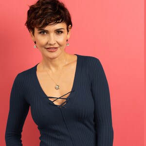 May include: A woman with short brown hair wearing a dark blue v-neck sweater with a gold necklace and matching earrings. The necklace has a round pendant with a black and white pattern.