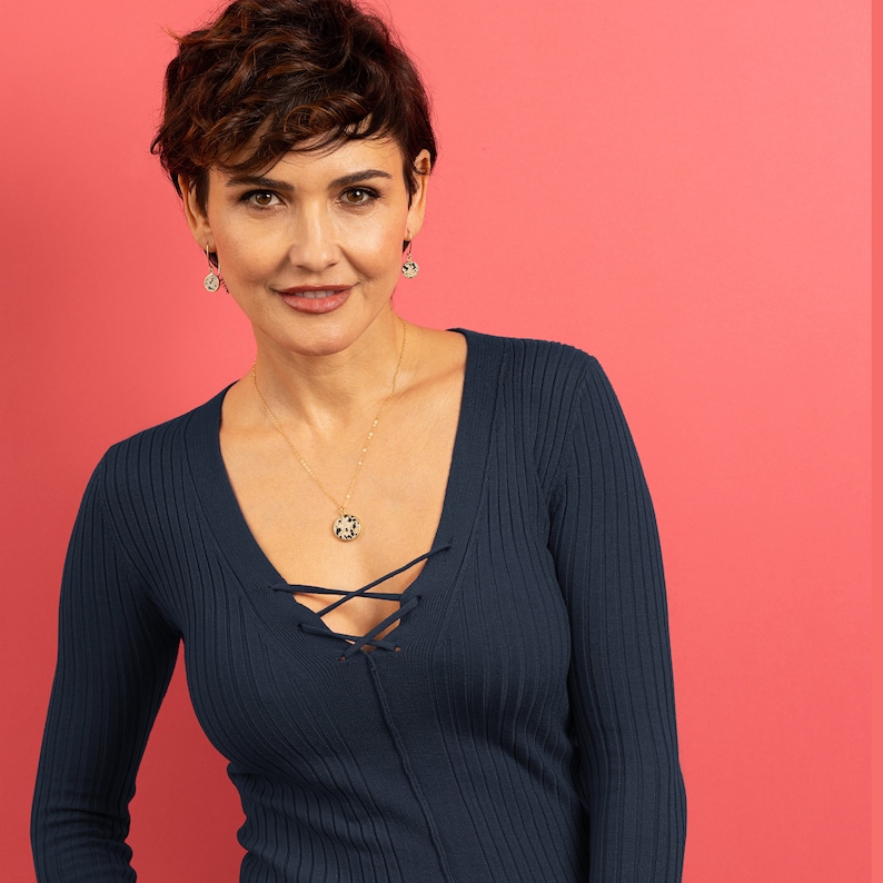 May include: A woman with short brown hair wearing a dark blue v-neck sweater with a gold necklace and matching earrings. The necklace has a round pendant with a black and white pattern.