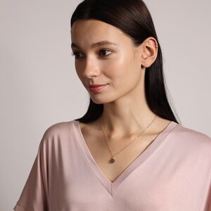 May include: A woman wearing a light pink v-neck shirt with a gold chain necklace and a small pendant. The pendant is a round, dark grey stone.