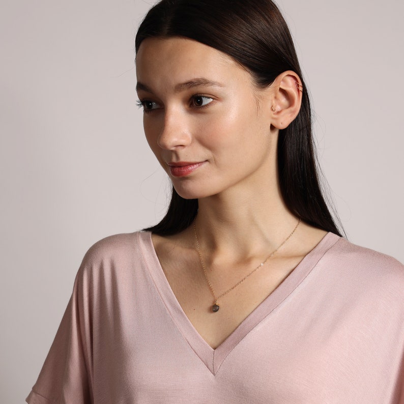 May include: A woman wearing a light pink v-neck shirt with a gold chain necklace and a small pendant. The pendant is a round, dark grey stone.