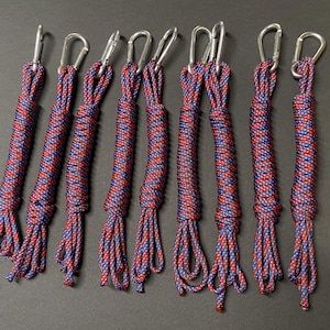 Könnte beinhalten: Acht rot-blaue geflochtene Seile mit silbernen Karabinerhaken. Die Seile sind eng gewickelt und haben eine dekorative Quaste am Ende. Der Hintergrund ist einfarbig dunkelgrau.