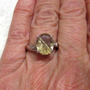 Peut inclure: Une bague argentée ornée d'une grande pierre précieuse jaune pâle de taille ovale, fixée par quatre griffes. L'anneau est agrémenté de petites pierres claires de taille ronde de chaque côté du sertissage central, offrant un aspect poli et brillant.