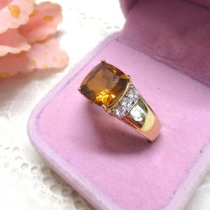 Può includere: Un anello in oro con una grande pietra preziosa color ambra a taglio cuscino al centro, affiancata da tre piccole pietre chiare a taglio rotondo su ciascun lato della fascia, presentato all'interno di un portagioie foderato in velluto viola chiaro.