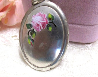 May include: Oval silver locket with a painted pink rose design. The locket has a loop at the top for a chain and is set against a soft pink background. A pink rose is visible in the background.