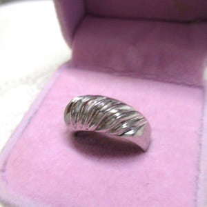 Op de afbeelding: Een zilverkleurige metalen ring met een brede, gewelfde band voorzien van een reeks diagonale, gegroefde ribbels over het oppervlak. De ring wordt gepresenteerd in een klein, open sieradendoosje met een lichtroze fluwelen voering.