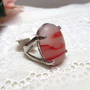 Könnte beinhalten: Ein silberfarbener Ring mit einem großen, ovalen, rosa Stein mit roten Adern. Der Ring liegt auf weißer Spitze, im Hintergrund eine rosa Blume. Ein einzigartiges Schmuckstück.
