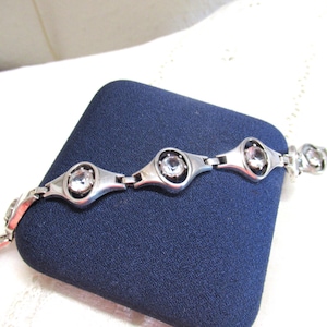 Puede incluir: Una pulsera plateada con eslabones ovalados, cada uno con una gema redonda y transparente. La pulsera se exhibe sobre una superficie azul oscuro, resaltando su diseño. Una joya.
