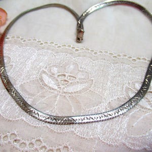 May include: A silver-toned herringbone chain necklace featuring a flat, flexible design with a subtle etched pattern across the surface. The necklace is shaped into a rounded loop and includes a small metal clasp mechanism at the end of the chain.