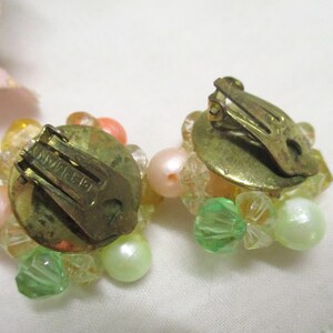 Może przedstawiać: Para vintage'owych kolczyk&oacute;w na klipsie z gromadą pastelowych r&oacute;żowych, zielonych i przezroczystych koralik&oacute;w. Kolczyki są oznaczone "SWAROVSKI" z tyłu.