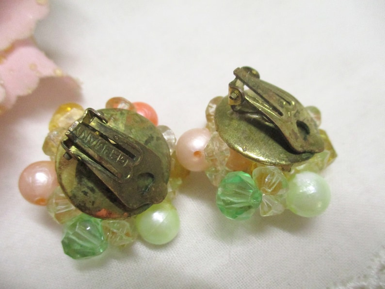 Może przedstawiać: Para vintage'owych kolczyk&oacute;w na klipsie z gromadą pastelowych r&oacute;żowych, zielonych i przezroczystych koralik&oacute;w. Kolczyki są oznaczone "SWAROVSKI" z tyłu.