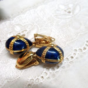 Può includere: Un paio di orecchini a clip color oro con pendenti a forma di uovo in smalto blu intenso. Ogni pendente è decorato con un motivo a croce dorato e testurizzato sulla superficie blu. Gli orecchini presentano un meccanismo a clip dorato lucido.