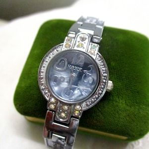 Könnte beinhalten: Eine silberne Uhr mit einem kristallbesetzten Band und Zifferblatt. Das Zifferblatt hat einen blauen Hintergrund mit silbernen römischen Ziffern als Stundenmarkierungen und einem Herzdesign. Die Uhr zeigt "HOUK" auf dem Zifferblatt.