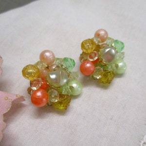 Może przedstawiać: Para vintage'owych klipsy z gromadą pastelowych zielonych, ż&oacute;łtych i pomarańczowych koralik&oacute;w. Kolczyki mają dużą, tęczową białą perłę w środku każdego skupiska.
