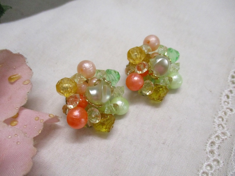 Może przedstawiać: Para vintage'owych klipsy z gromadą pastelowych zielonych, ż&oacute;łtych i pomarańczowych koralik&oacute;w. Kolczyki mają dużą, tęczową białą perłę w środku każdego skupiska.