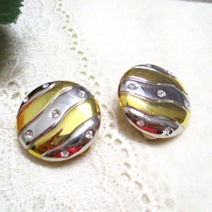 May include: Pair of round clip-on earrings. Each earring has a gold, silver, and gold striped design with small, clear rhinestones. The earrings have a shiny, metallic finish and a classic, elegant style.
