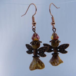 Puede incluir: Un par de pendientes hechos a mano con un diseño de hada. Los pendientes tienen ganchos de color cobre y están adornados con cuentas en tonos amarillos, rojos y marrones. Las alas y la falda del hada tienen un acabado texturizado.