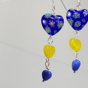May include: A pair of dangle earrings with blue and yellow heart-shaped beads. The top heart bead is blue with a white floral pattern. The bottom heart bead is yellow. The earrings are silver.
