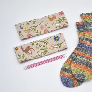 May include: A pair of hand-knitted socks with horizontal stripes in shades of blue, orange, green, and gray. Two fabric needle cases with floral and deer patterns are also visible, along with a set of pink knitting needles.