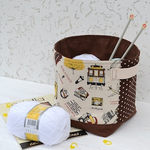 May include: A brown and white fabric storage bin with a polka dot pattern and a handle. The bin is filled with white yarn and knitting needles. The bin has a design of a trolley car and the text "Bon Voyage" on it.